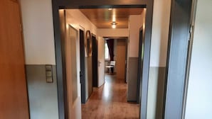 A narrow hallway with wooden flooring, leading to a room with a window and a chair.