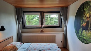 A bedroom with a wooden ceiling, a bed with floral bedding, a radiator, a window with curtains, and a wall painting of a deer.
