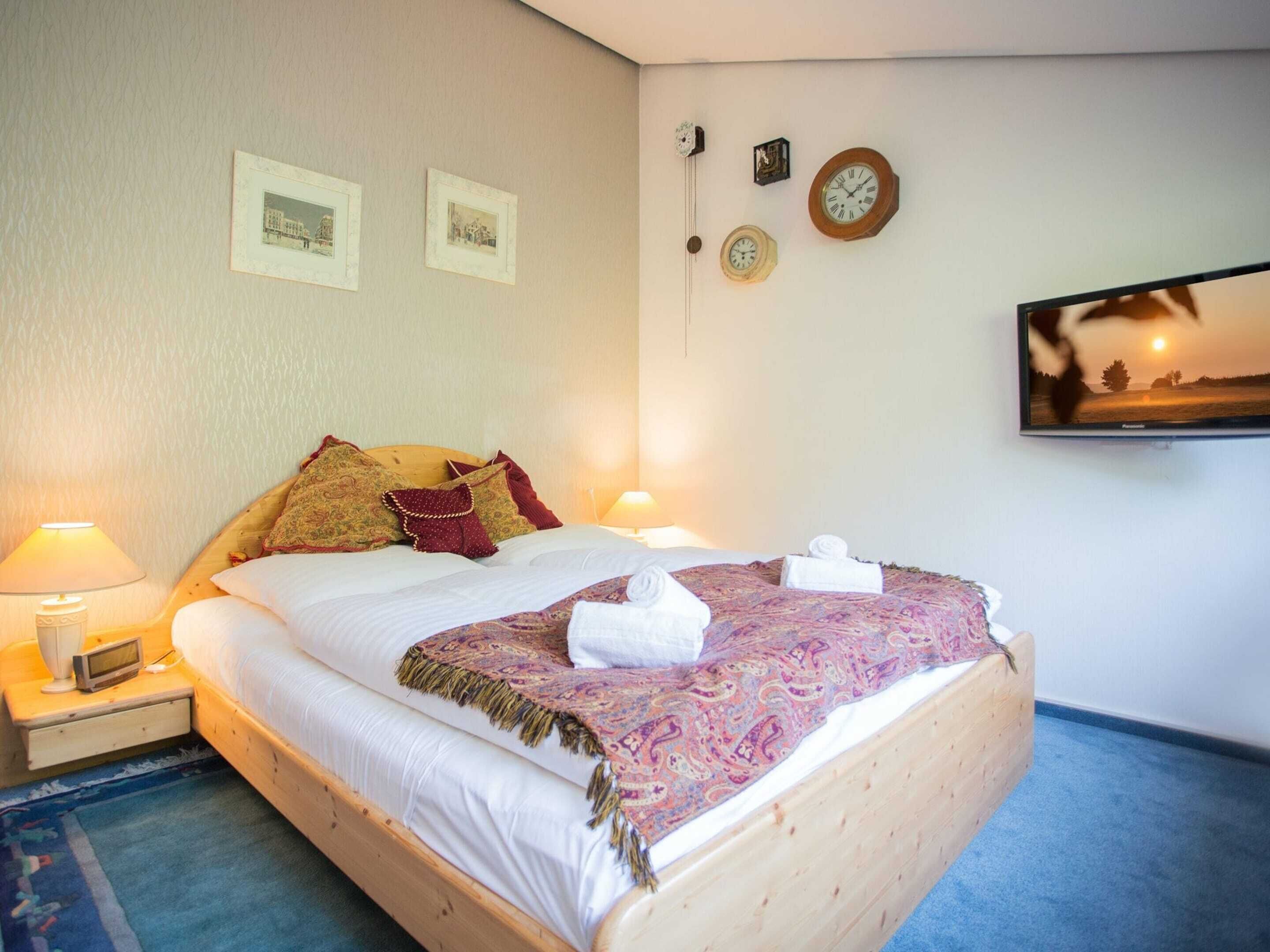 Een net opgemaakt bed met een gebogen deken, twee bedlampen, een klok en een televisie aan de muur.