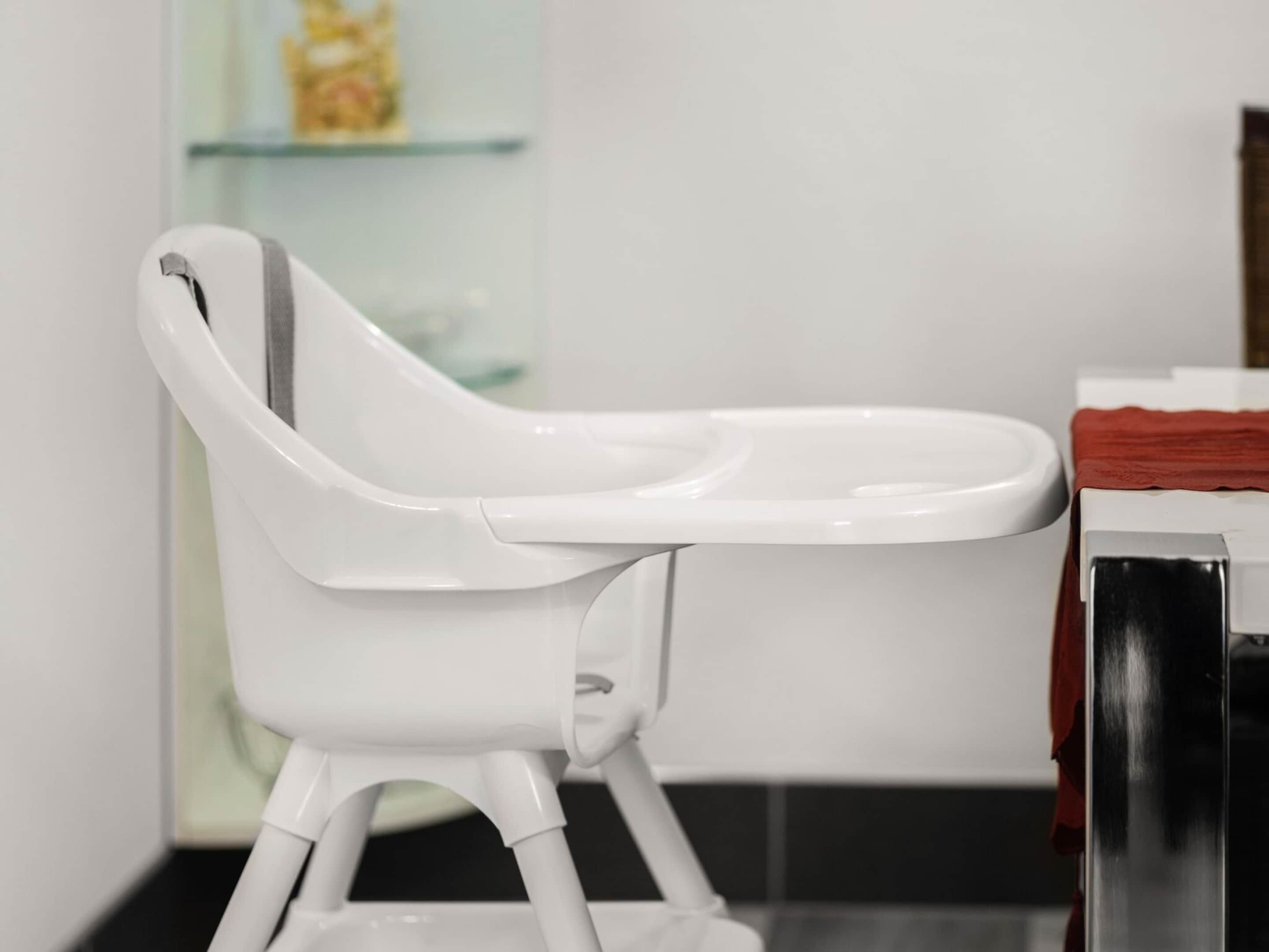 A white high chair with a tray, positioned in a room with a glass cabinet and a red cloth on a table.