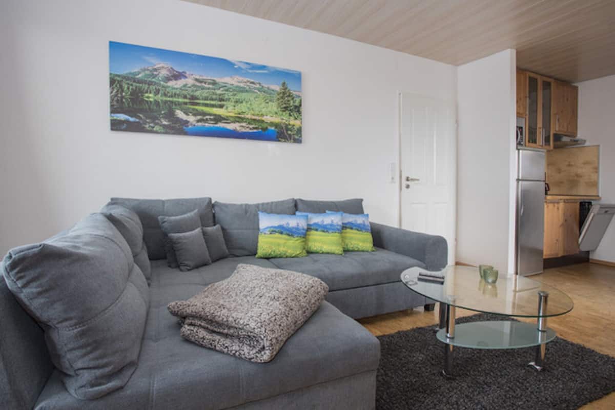 Uma sala de estar moderna com um sofá cinzento, uma mesa de centro de vidro e um quadro de uma paisagem na parede.