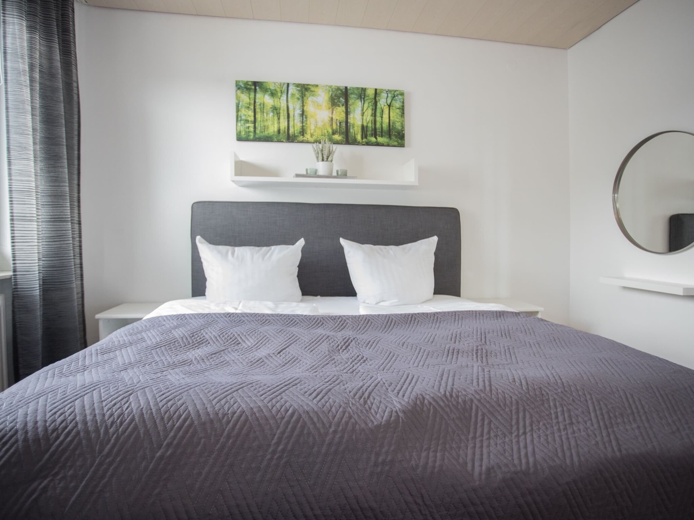 A modern hotel room with a large bed, a headboard, a bedside table, a mirror, and a wall-mounted shelf with a painting.