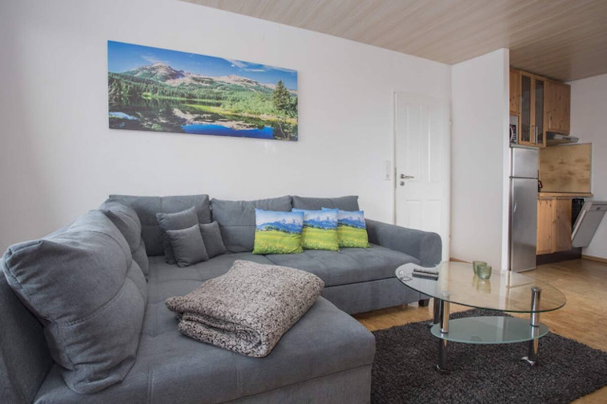 A modern living room with a grey sofa, a glass coffee table, and a painting of a landscape on the wall.