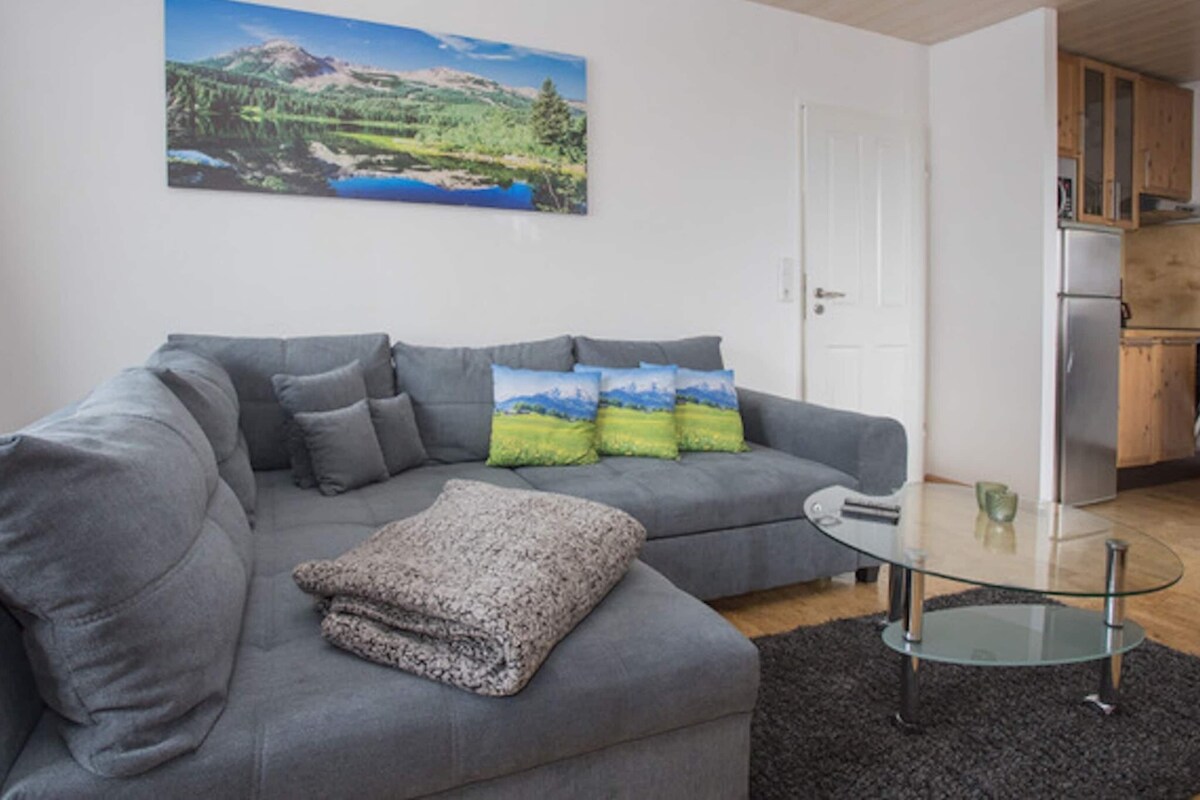 A modern living room with a grey sectional sofa, a glass coffee table, and a painting of a landscape on the wall.