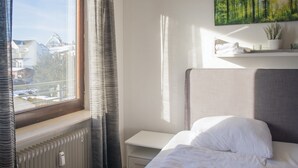 A hotel room with a bed, a nightstand, a window with curtains, a radiator, and a wall-mounted shelf with a plant and a painting. - Wohnung in Winterberg mit Balkon in Pistennahe (Winterberg)