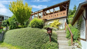 Une maison à deux étages dotée d’un balcon en bois, d’un escalier menant à l’entrée et d’un jardin bien entretenu.