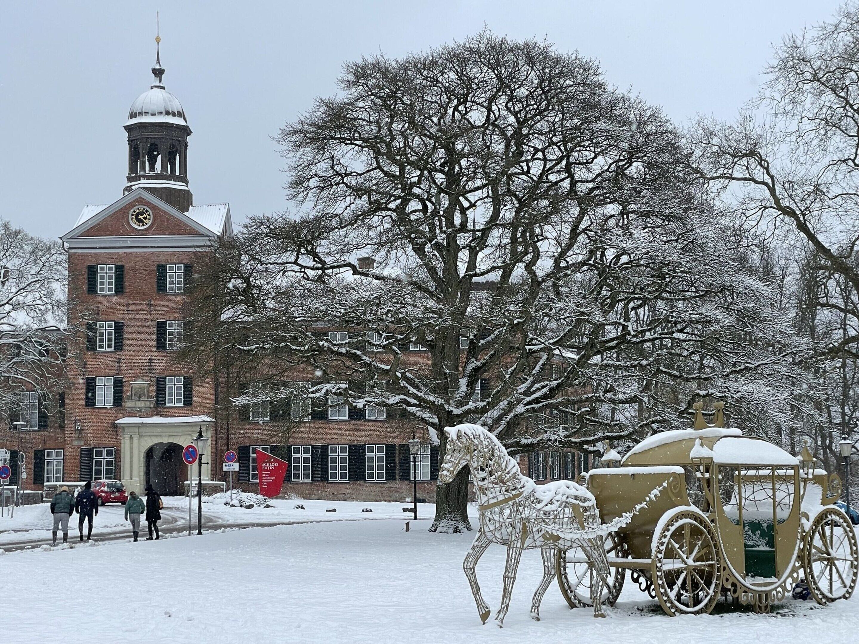A snow-covered scene with a historic building, a horse-drawn carriage, and bare trees.