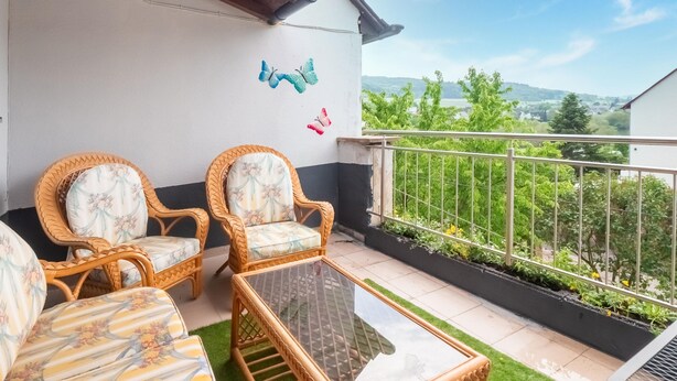 Varanda com móveis de vime, mesa de vidro e vista para áreas verdes e edifícios ao longe.