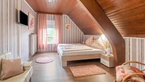 Um quarto de sótão acolhedor, com teto inclinado, uma cama de casal, um pequeno espaço de estar com uma poltrona e vista para o verde lá fora.