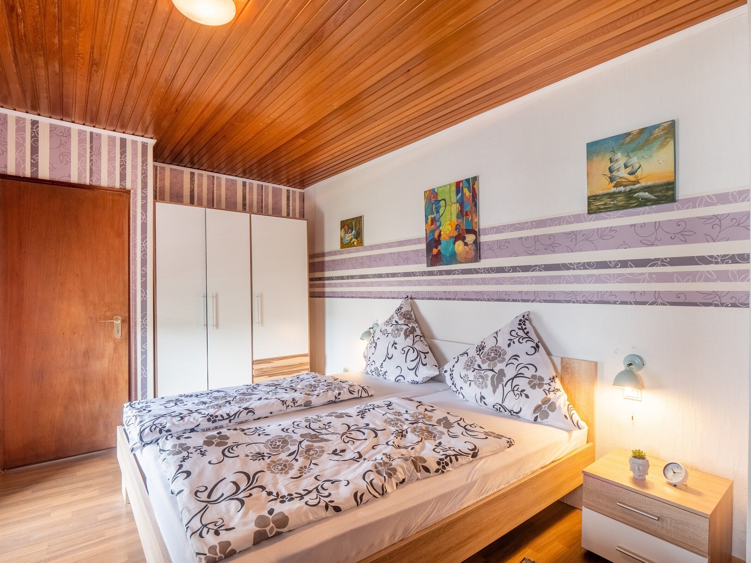 A bedroom with a wooden ceiling, a bed with floral bedding, a nightstand with a lamp, and artwork on the wall.