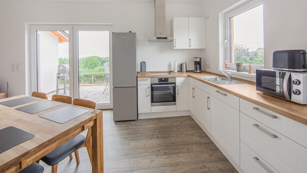 A modern kitchen with white cabinets, a built-in oven, a microwave, and a coffee maker. There is a dining table with chairs and a sliding glass door leading to an outdoor area. - Ferienwohnung Eifelruhe in Filz (Filz)