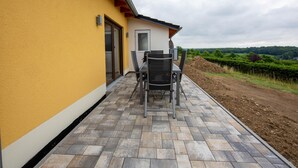 A patio with a table and chairs, a building with yellow walls, and a view of a green landscape. - Ferienwohnung Eifelruhe in Filz (Filz)