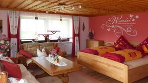 Ein gemütliches Zimmer mit einem hölzernen Bett, einem Holztisch und einem Sofa. Ein Fenster mit Vorhängen und eine Wand mit einem dekorativen Schild sind ebenfalls vorhanden.