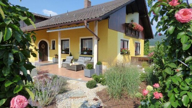 Ein gelbes Haus mit Terrasse, umgeben von Grünpflanzen und Blumen.