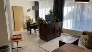 Ein Wohnzimmer mit Holzesstisch, einer Couch, einem Fernseher auf einer Holzkommode, einer Gitarre an der Wand und einem Fenster mit Vorhängen.
