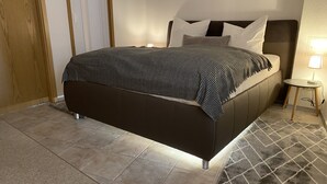 Ein modernes Schlafzimmer mit einem dunkel gepolsterten Bett, einer gemusterten Tagesdecke und einem kleinen Nachttischchen mit einer Lampe.