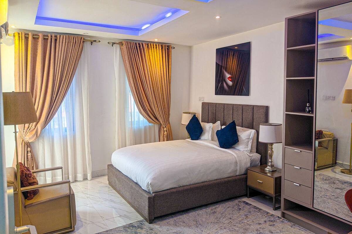 A modern hotel room with a large bed, bedside tables, a wardrobe, and a ceiling with blue lighting.