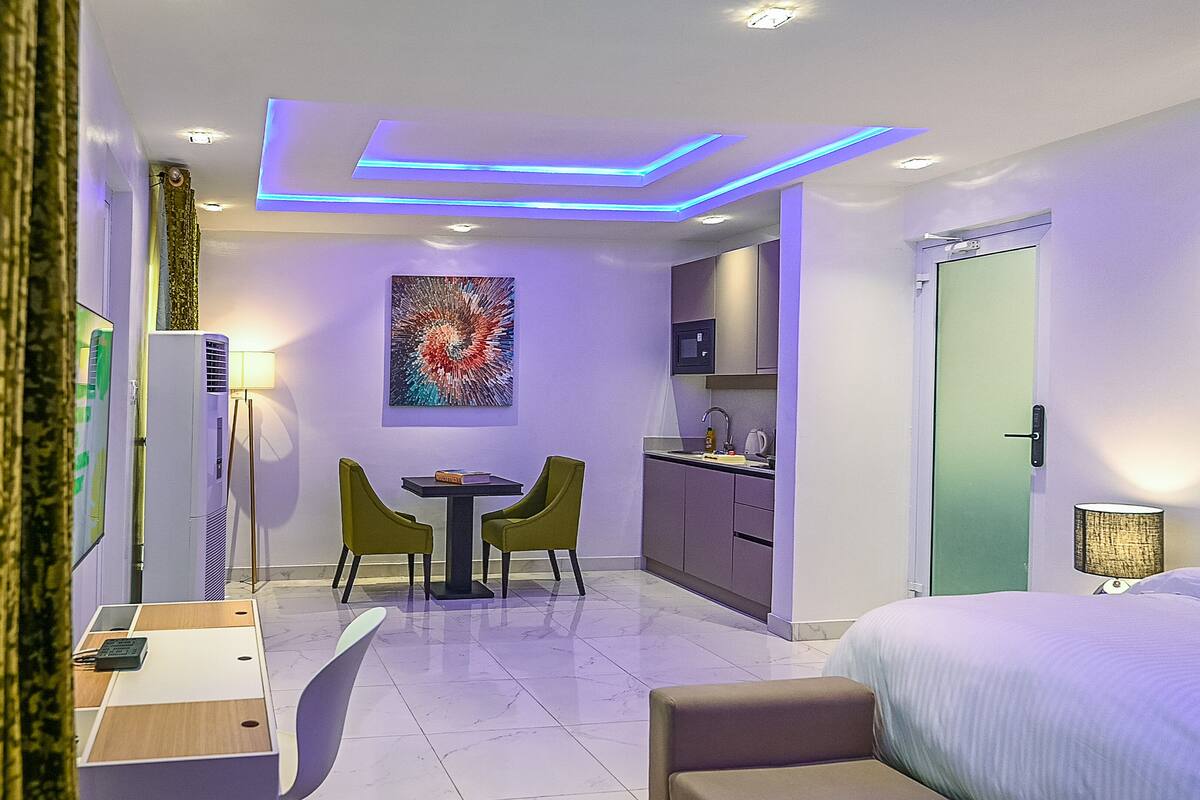 A modern hotel room with a bed, a desk, a chair, a small table, a microwave, a sink, and a door.