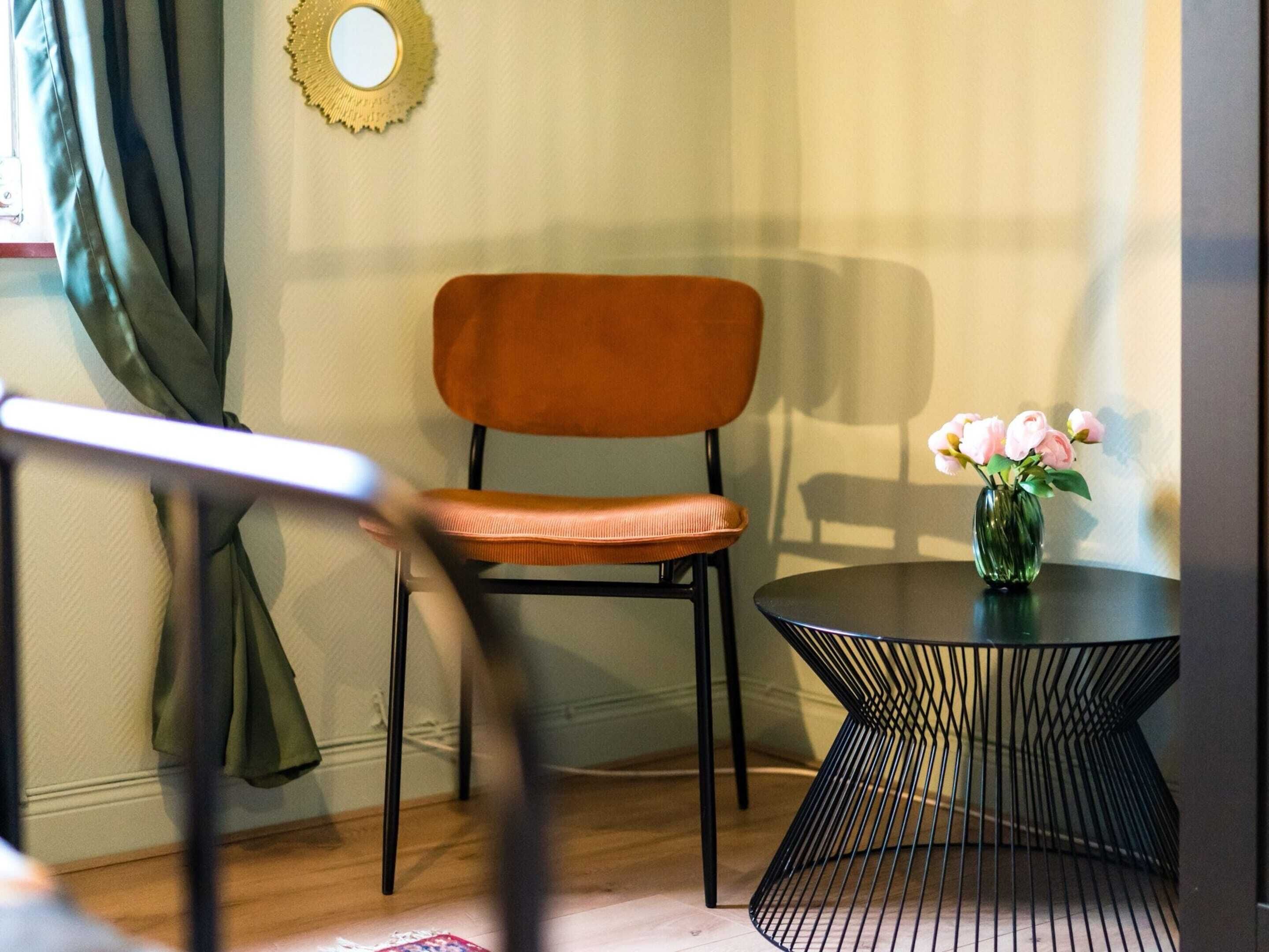 A room with a chair, a small table with a vase of flowers, and a mirror on the wall.