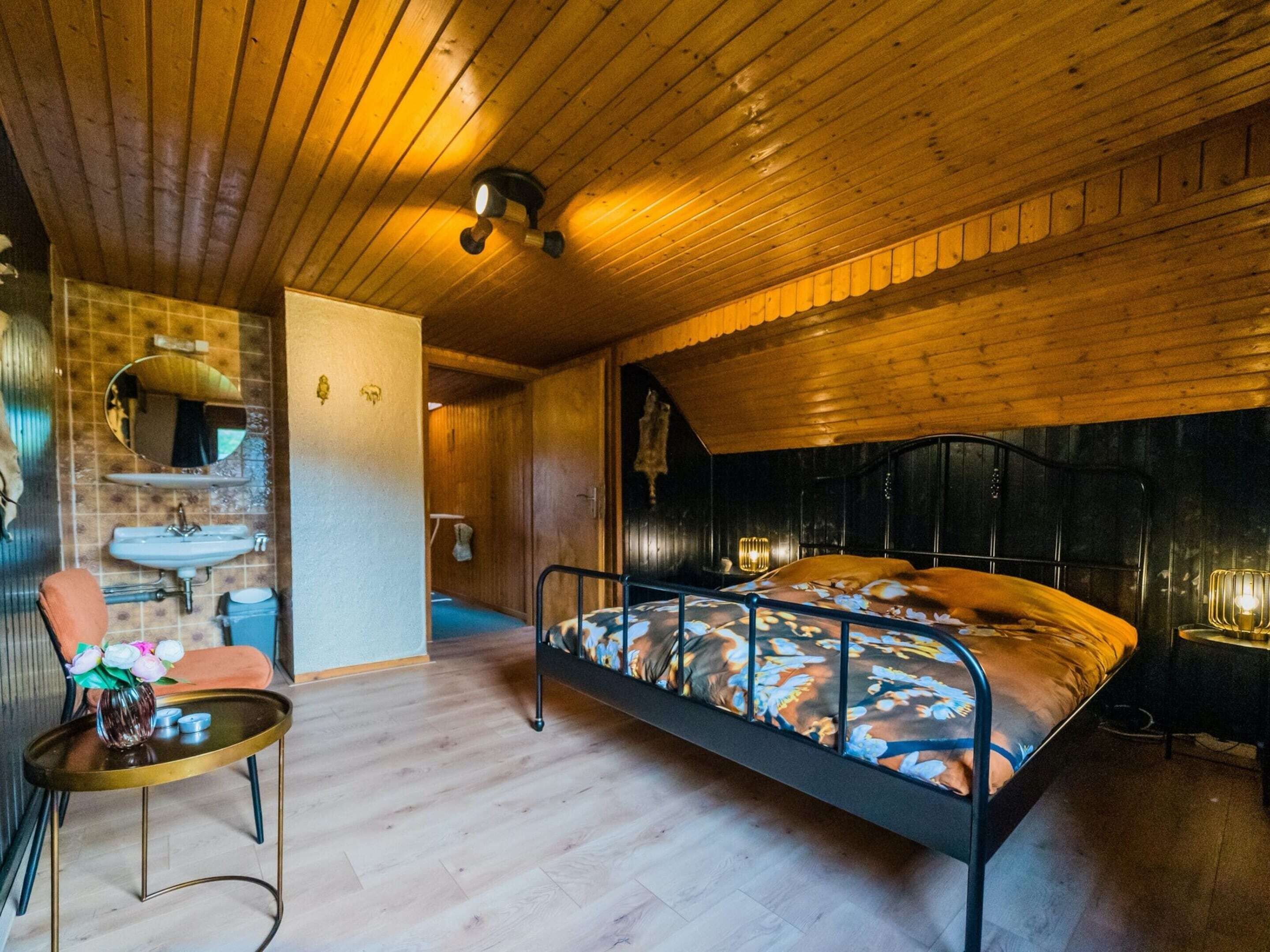 Camera da letto in una baita di legno, con un letto, un piccolo tavolino con un vaso, un lavandino e uno specchio.