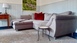 A modern living room with a grey sectional sofa, a small round table, a lamp, and a painting of a landscape. - Holiday Home With Natural Garden (Kaltennordheim)