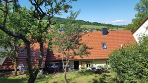 A house with a red-tiled roof, a small window, and a garden with a tree and a chair. - Holiday Home With Natural Garden (Kaltennordheim)