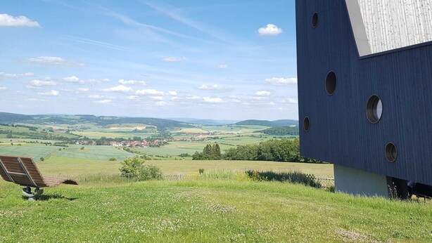 A modern building with a sloped roof and circular windows, overlooking a green landscape with fields and distant houses. - Holiday Home With Natural Garden (Kaltennordheim)