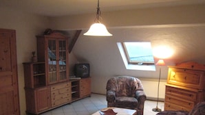A cozy living room with a wooden cabinet, a television, a patterned armchair, a coffee table, and a lamp.