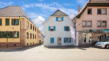 Eine Reihe von Gebäuden, mit einem zentralen weißen Gebäude, das eine Tür und Fenster hat, umrahmt von Gebäuden mit grünen Fensterläden und einem rosafarbenen Gebäude mit einem Schild.