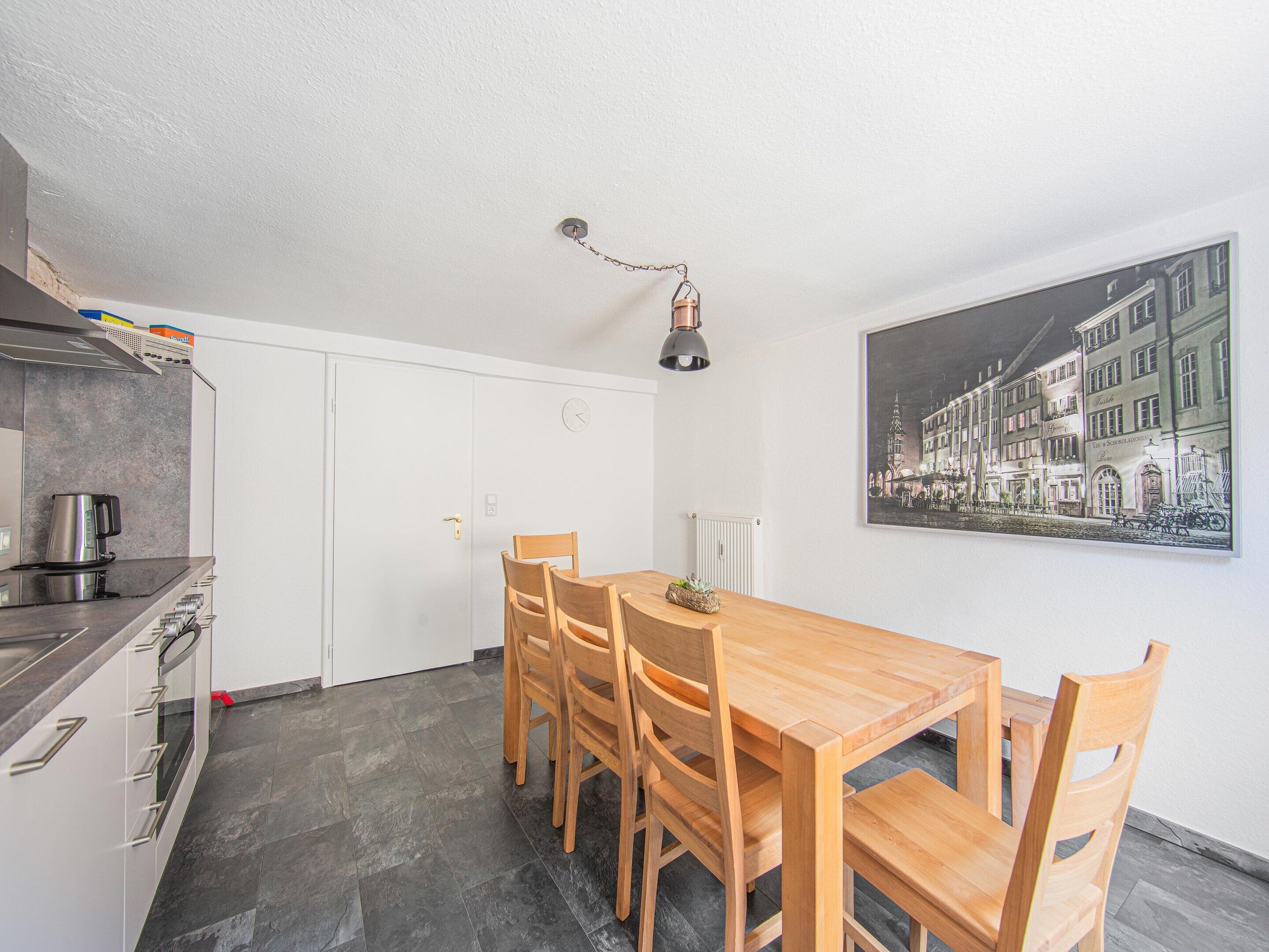 A compact kitchen with a dining area, featuring a wooden table and chairs, a coffee machine, and a wall-mounted clock.
