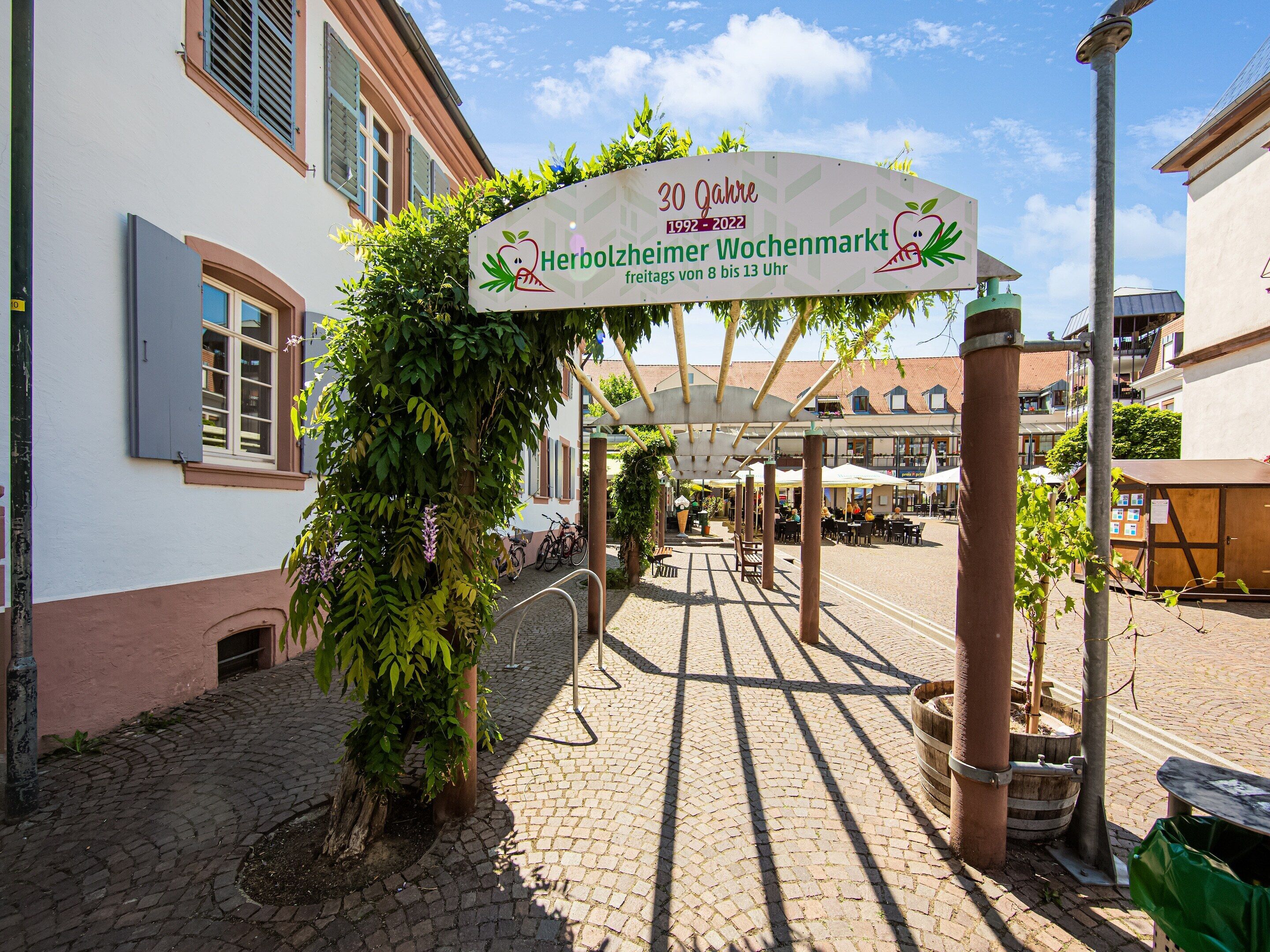 Ein Kopfsteinpflasterhof mit überdachter Sitzgelegenheit, einem Schild zur 30-jährigen Wiederkehr und einem Gebäude mit weißer Fassade und Fensterläden.