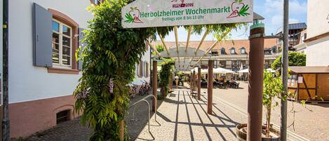 Ein Kopfsteinpflasterhof mit überdachter Sitzgelegenheit, einem Schild zur 30-jährigen Wiederkehr und einem Gebäude mit weißer Fassade und Fensterläden.