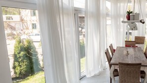 A dining area with a wooden table and wicker chairs, large windows with white curtains, and a hanging plant. - Luxurious Holiday Home With a Nice Private Garden (Anschau)