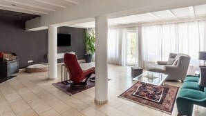 A spacious living room with a modern design, featuring a red chair, a grey sofa, and a teal sofa. There is a large window with sheer curtains, a flat-screen TV mounted on the wall, and a potted plant. - Luxurious Holiday Home With a Nice Private Garden (Anschau)