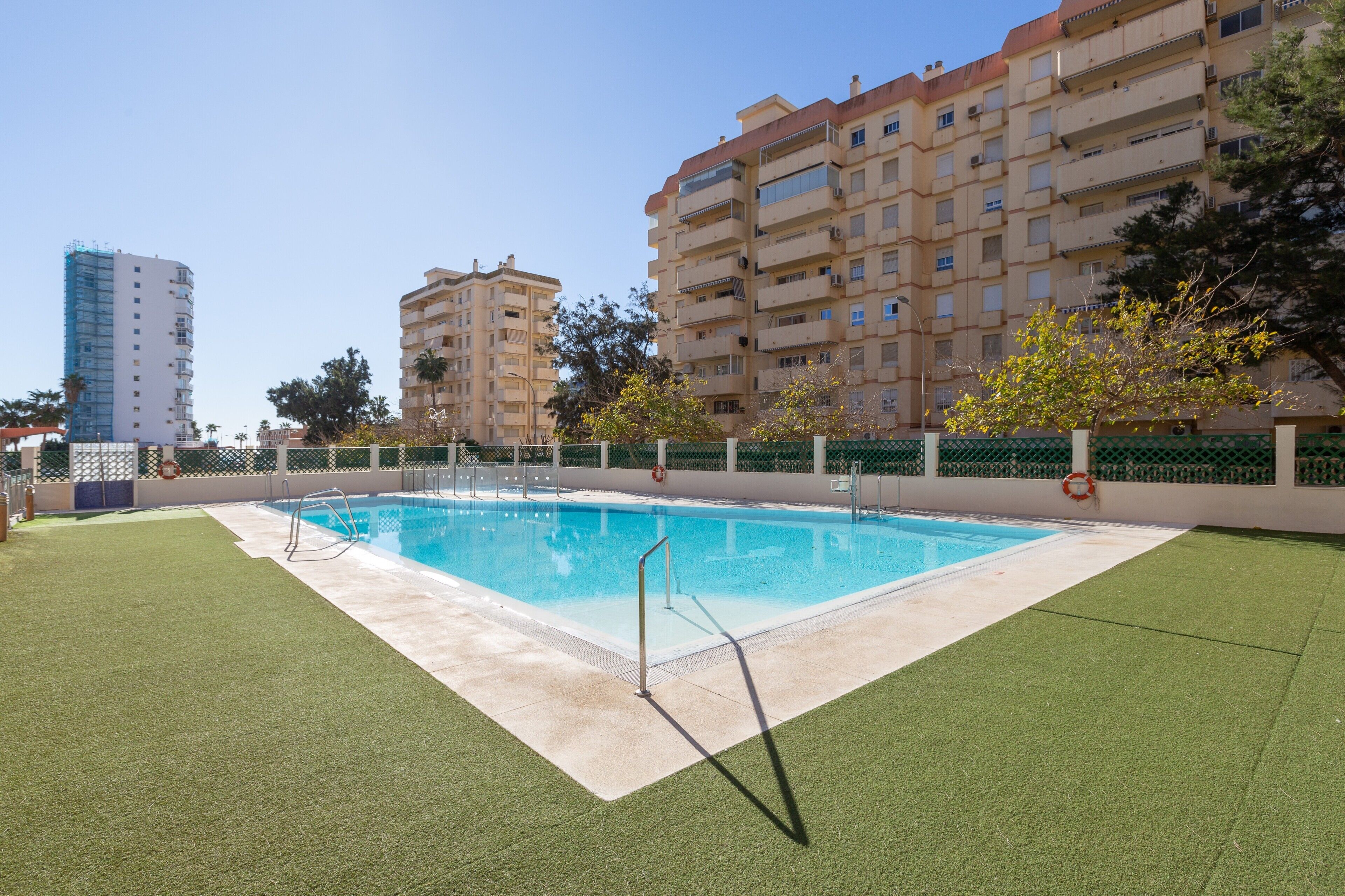 Una piscina con una silla de salvavidas, rodeada de césped artificial, y edificios de apartamentos al fondo.