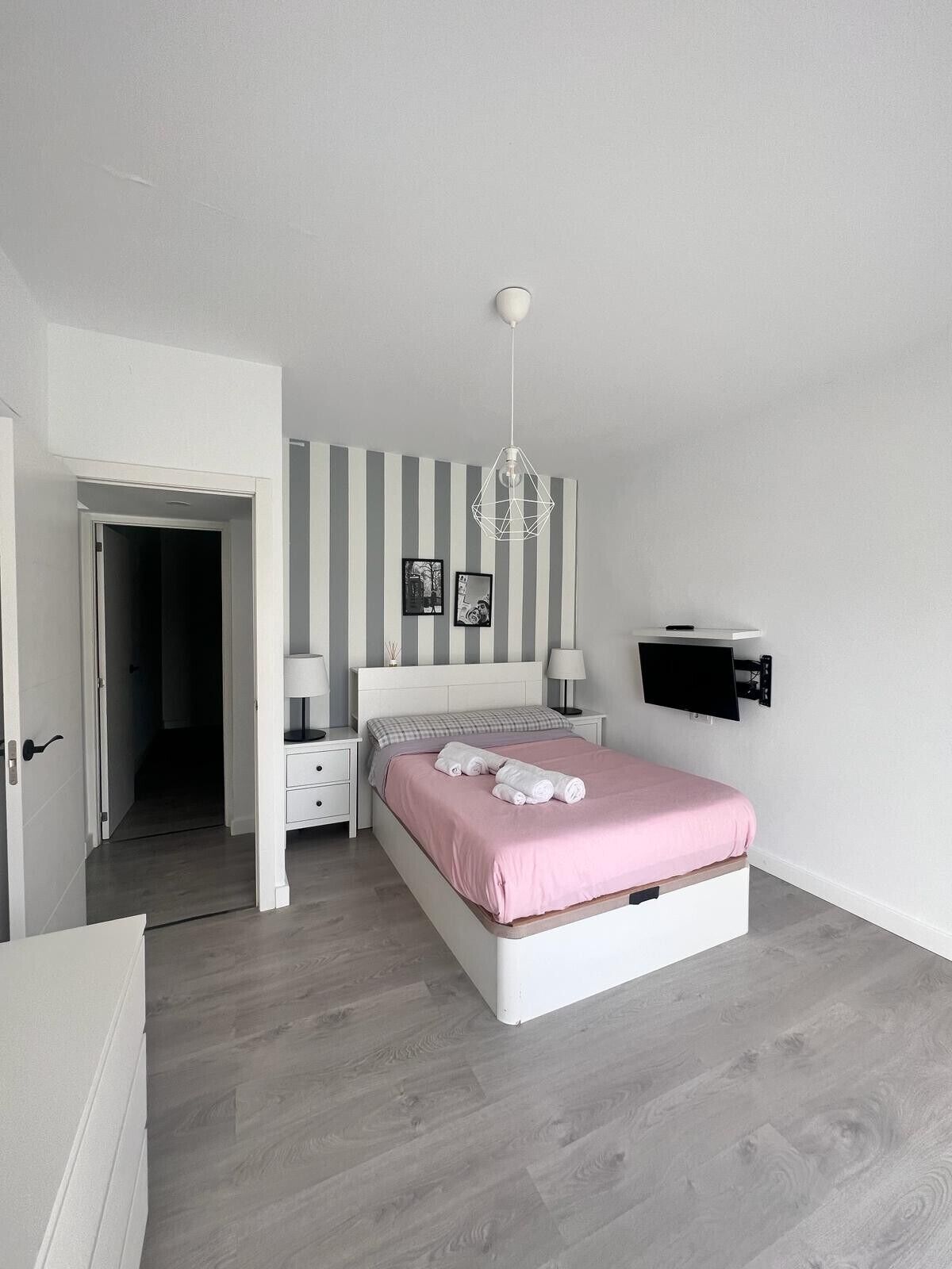 Un dormitorio con cama, mesitas de noche, lámpara colgante, televisor instalado en la pared y una puerta que da a otra habitación.