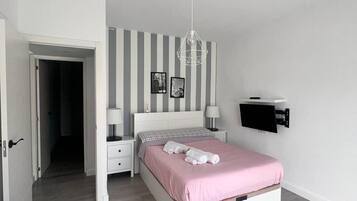Un dormitorio con cama, mesitas de noche, lámpara colgante, televisor instalado en la pared y una puerta que da a otra habitación.