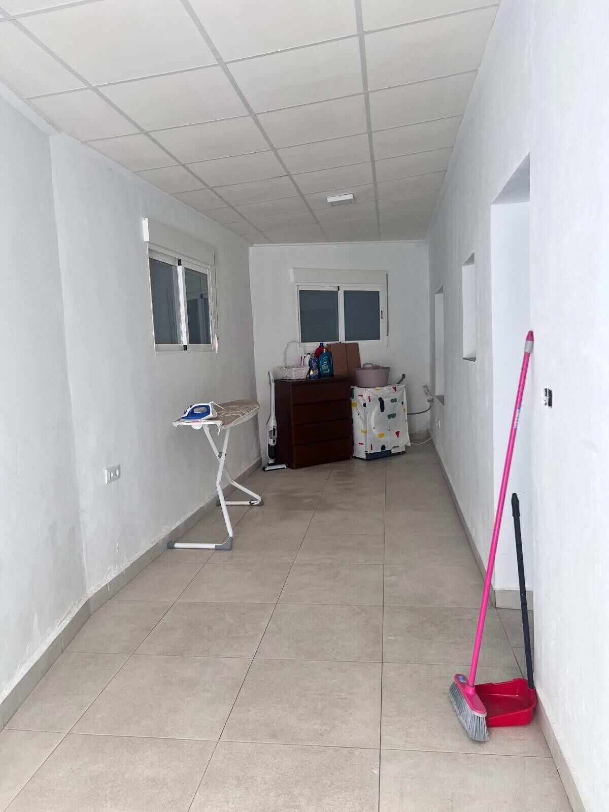 A room with a tiled floor, a desk with a chair, a window, and a broom leaning against the wall.