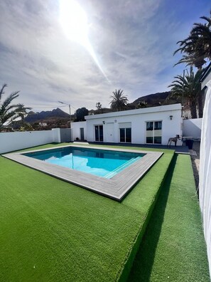 Una piscina rettangolare con un prato sintetico verde circostante, un edificio bianco con una porta e finestre, e palme sullo sfondo.