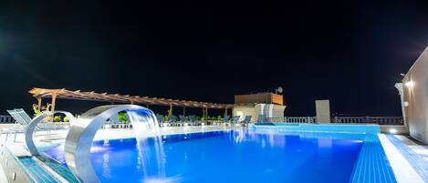 Una piscina nocturna con una fuente circular, sillas de descanso y un área de estar cubierta.