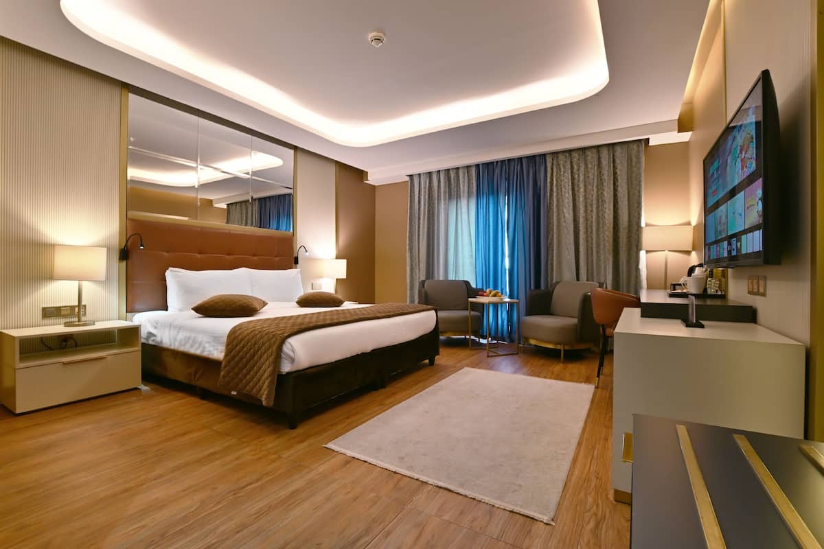 Una habitación de hotel moderna con una cama grande, mesitas de noche, un escritorio, una silla, un televisor y un ventanal con cortinas.