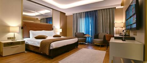 A modern hotel room with a large bed, bedside tables, a desk, a chair, a TV, and a large window with curtains.