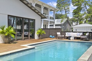 Ein moderner Poolbereich im Freien mit Holzterrasse, einer Sitzecke mit Sofa und Sesseln sowie einem großen Fenster mit Blick auf den Pool.