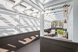 Alpendre coberto com deck de madeira, pérgola branca e área de estar com almofadas e um vaso de planta.