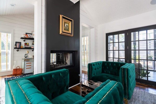 Un salon avec une cheminée, des fauteuils à revêtement vert, une table basse et une bibliothèque.