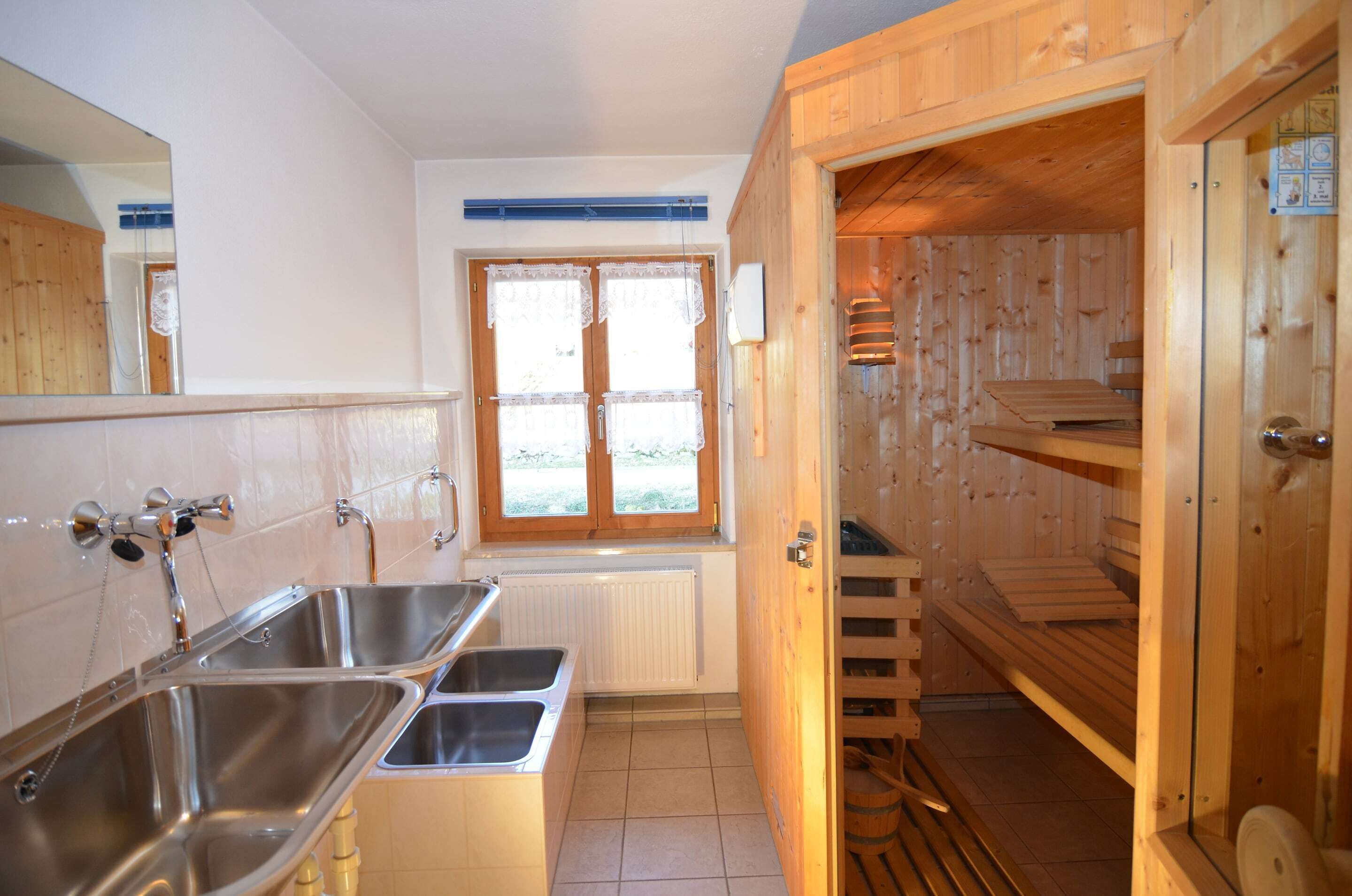 A compact kitchen with two stainless steel sinks, a window with lace curtains, and a wooden sauna visible through an open door.
