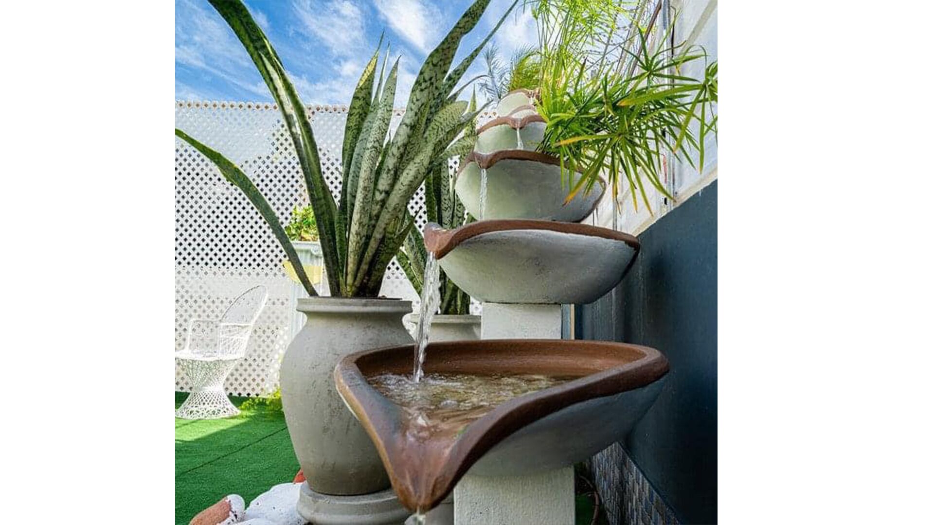 A tiered water feature with multiple bowls, surrounded by plants and a white fence.