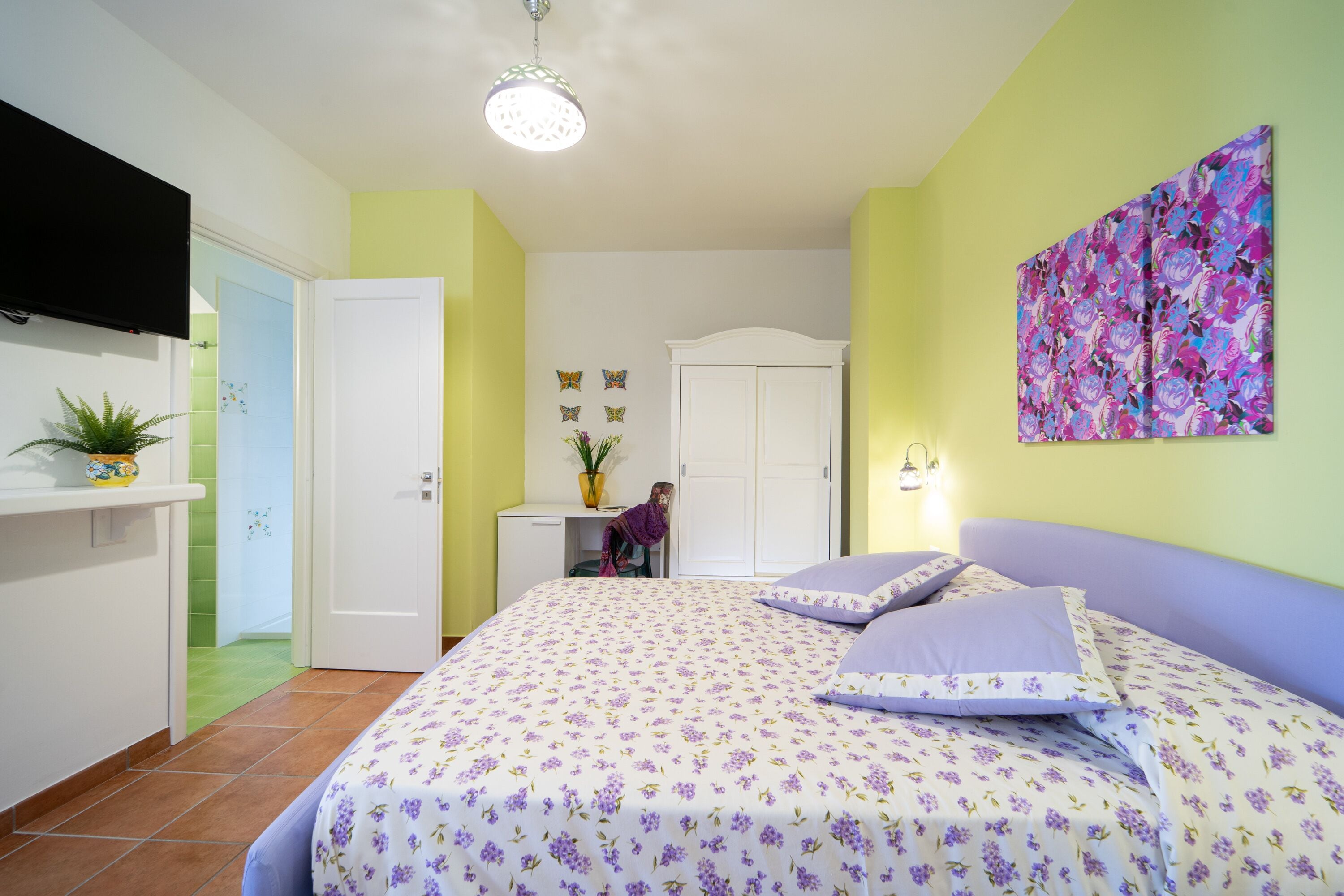 A bedroom with a bed, a TV mounted on the wall, a desk with a chair, and a door leading to another room.