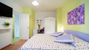 A bedroom with a bed, a TV mounted on the wall, a desk with a chair, and a door leading to another room.