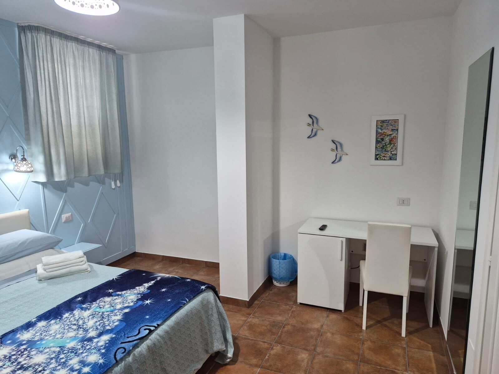 A bedroom with a bed, a desk, a chair, a small trash bin, and a wall-mounted light fixture.